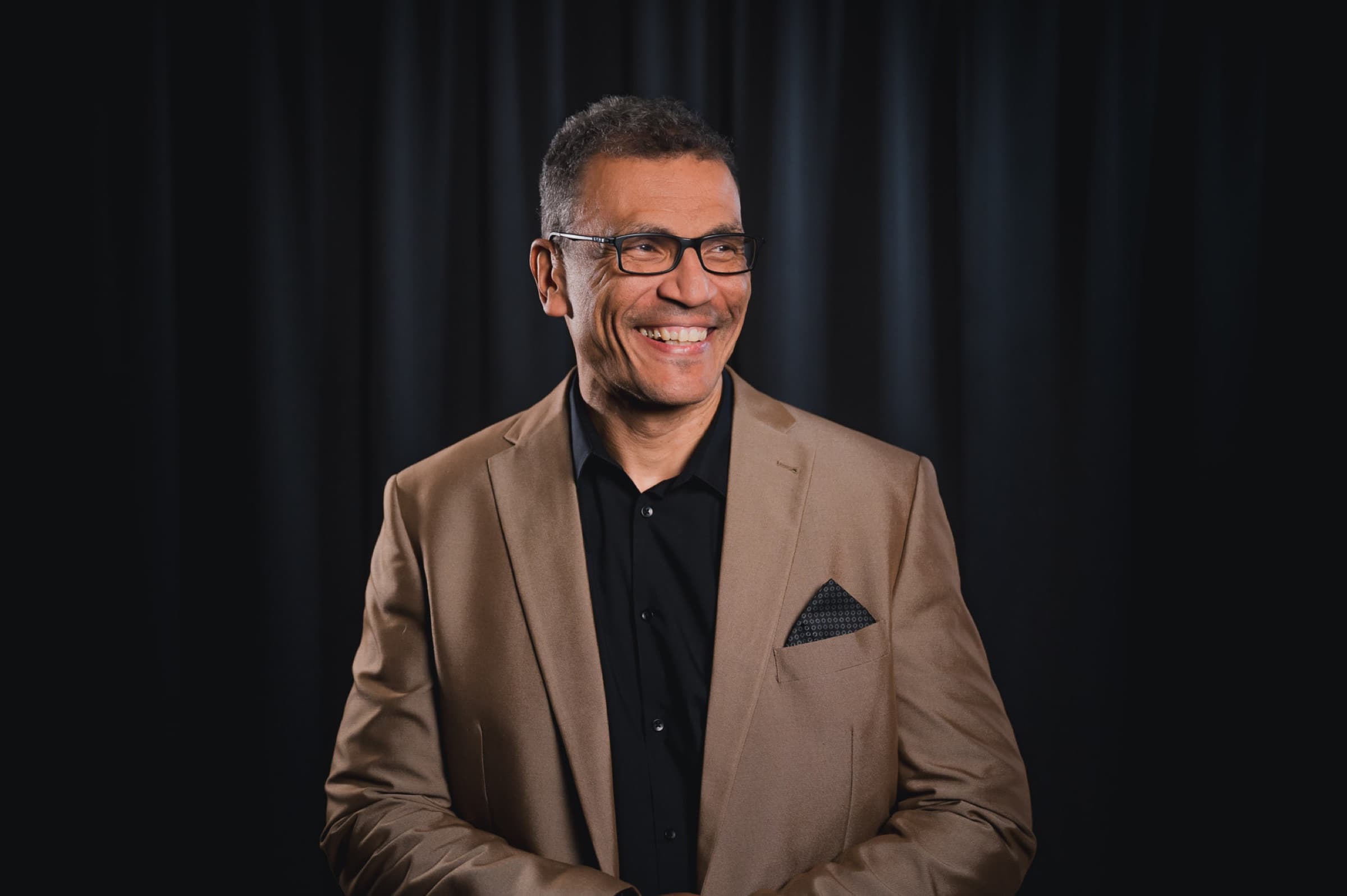 Dr. med. Orell Mielke, lachend, im hellbraunen Sakko vor dunklem Vorhang.