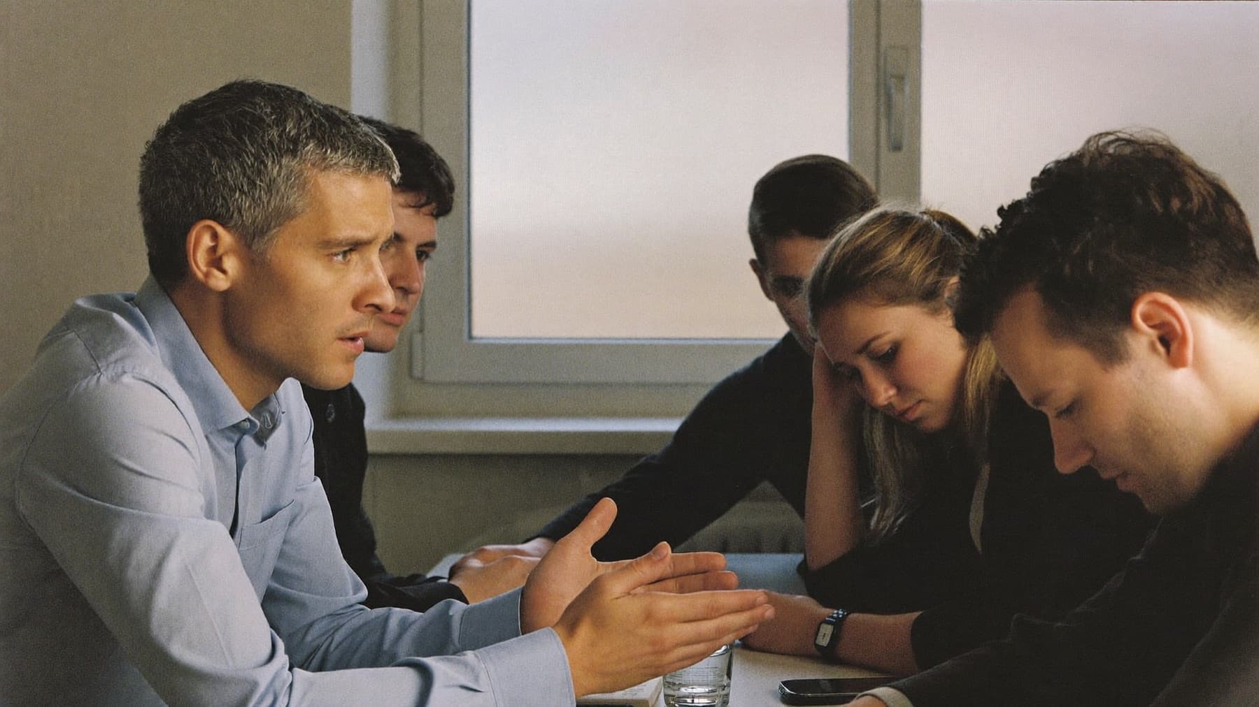 Fuenf Menschen an einem Konferenztisch: einer spricht, zwei sitzen mit gesenktem Blick da.